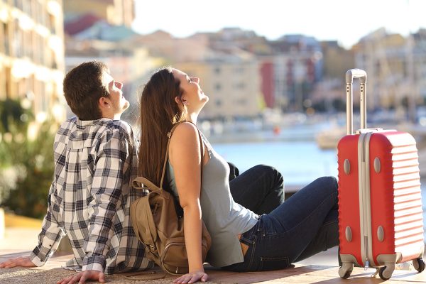 Side view of a couple of 2 tourists with a suitcase sitting relaxing and enjoying vacations in a colorful promenade. Tourism concept